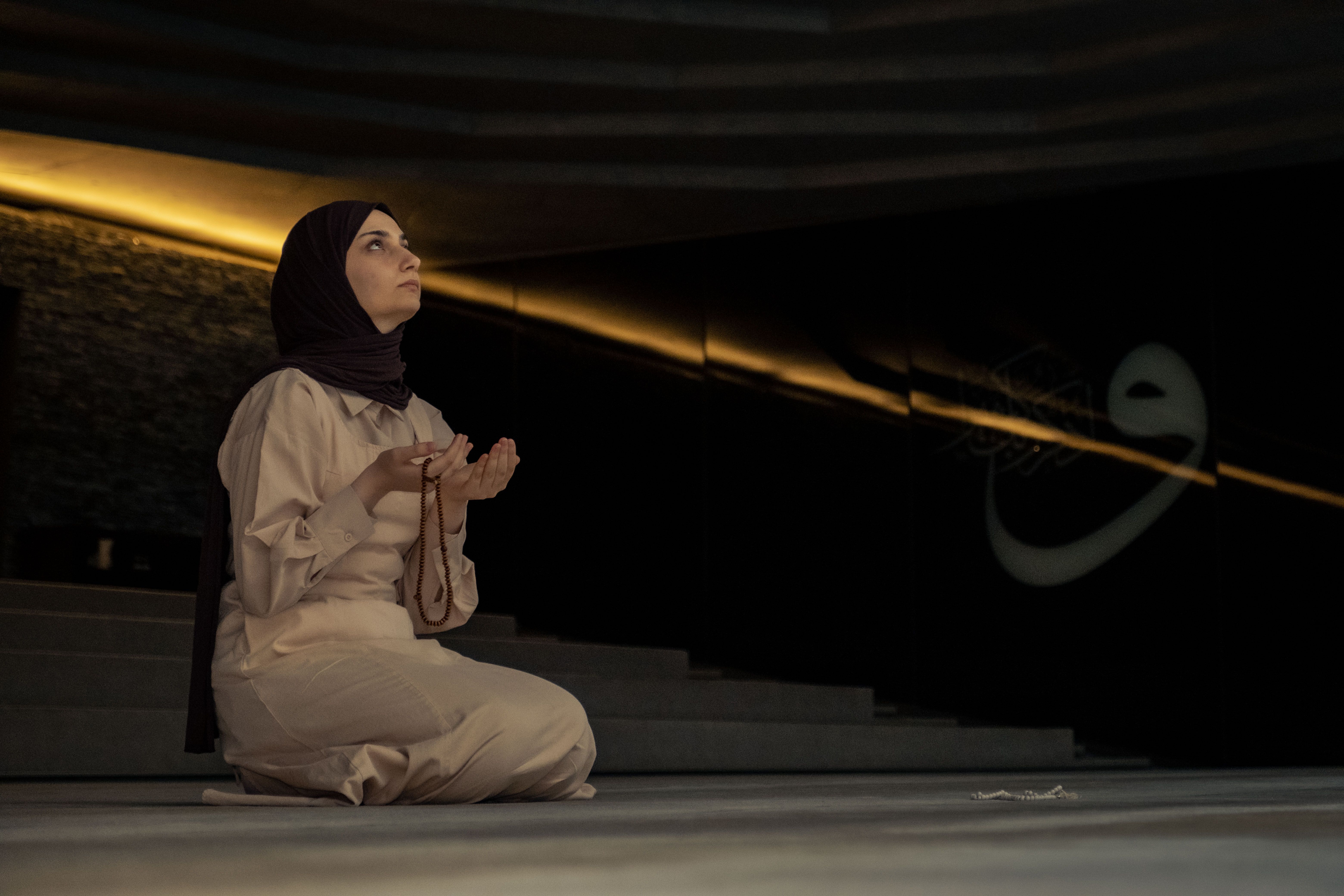 A woman in a hijab kneeling and praying inside a dimly lit mosque.