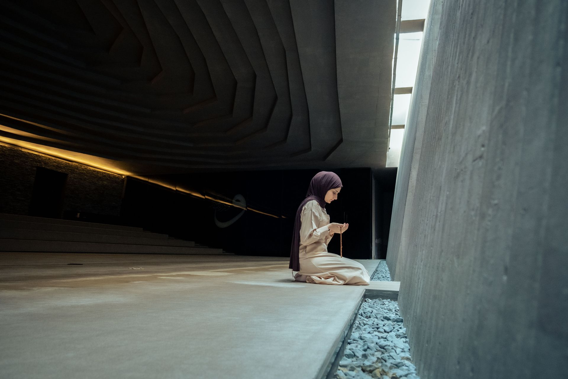 A young Middle Eastern woman in a hijab praying indoors, holding prayer beads.