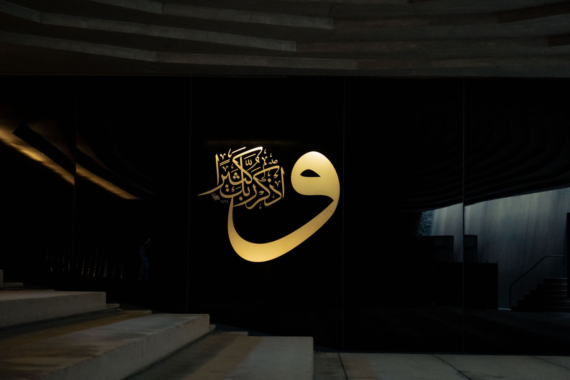 Elegant Islamic calligraphy in gold on a modern black wall, featuring architectural elements and stairs