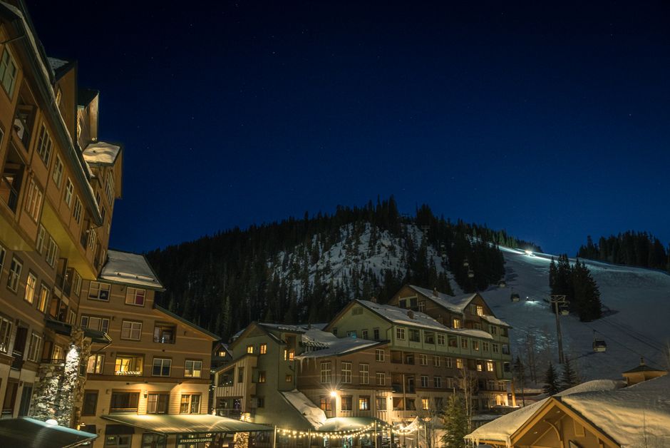 Illuminated ski resort town at night with snowy mountains under a starry sky in winter.