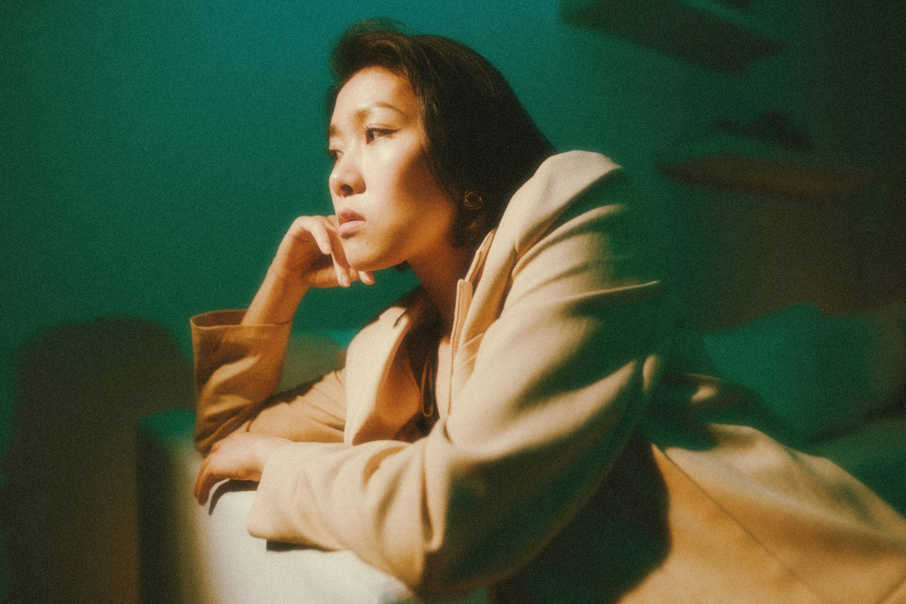 A woman in a beige jacket poses thoughtfully under green lighting.