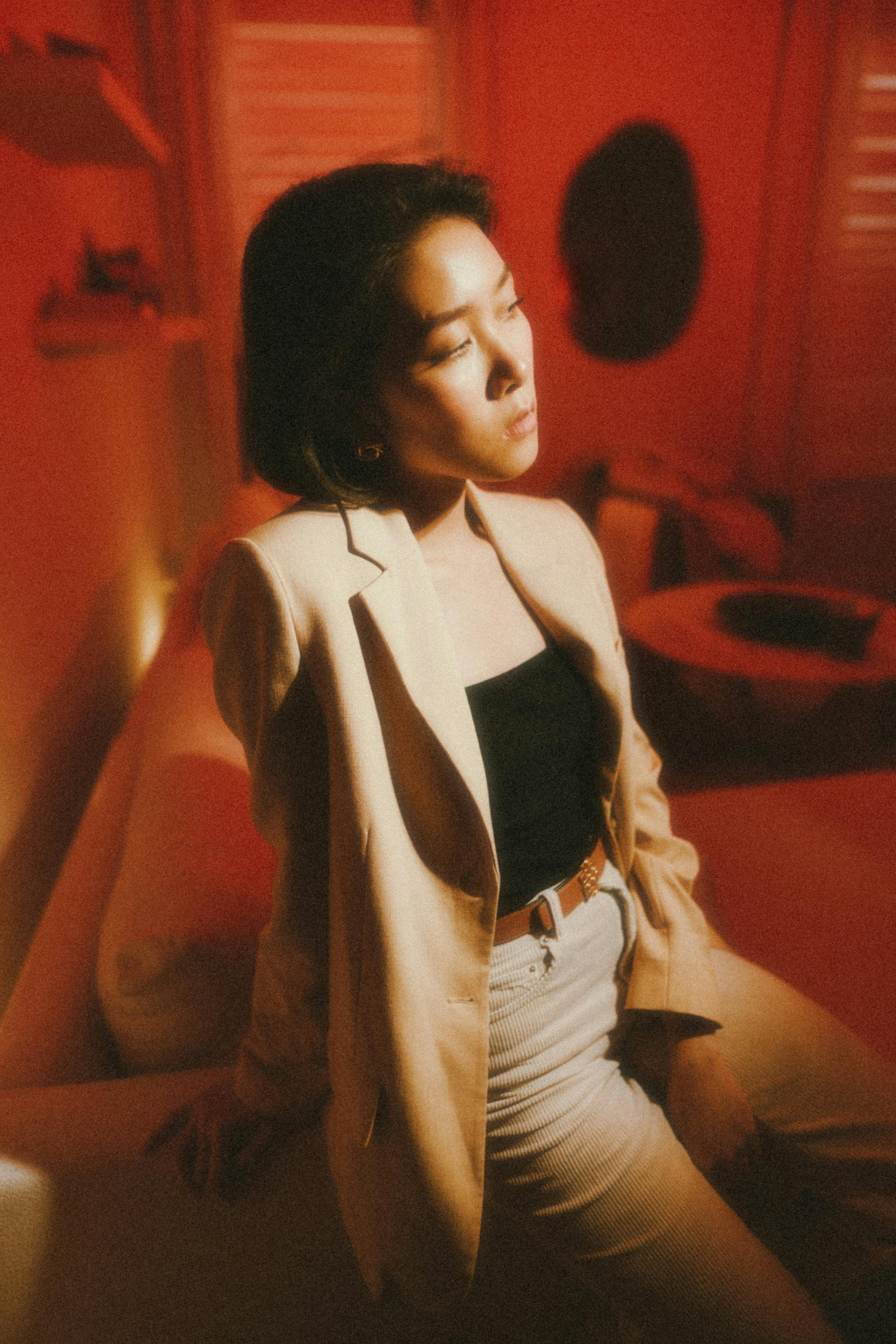 Portrait of an adult woman posing indoors with red lighting, wearing a beige jacket and black top.