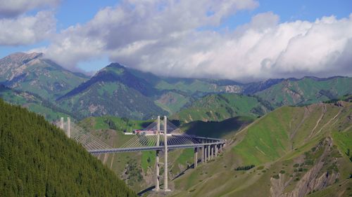 Free Scenic view of bridge spanning lush hills and Tianshan mountains under a cloudy sky. Stock Photo