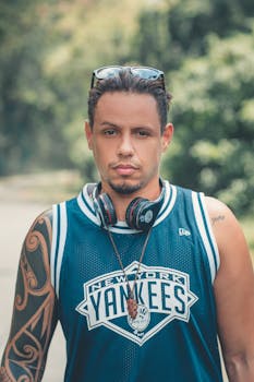 一个身穿纽约洋基队球衣、戴着耳机、身上有纹身的男人，神情严肃地站在户外