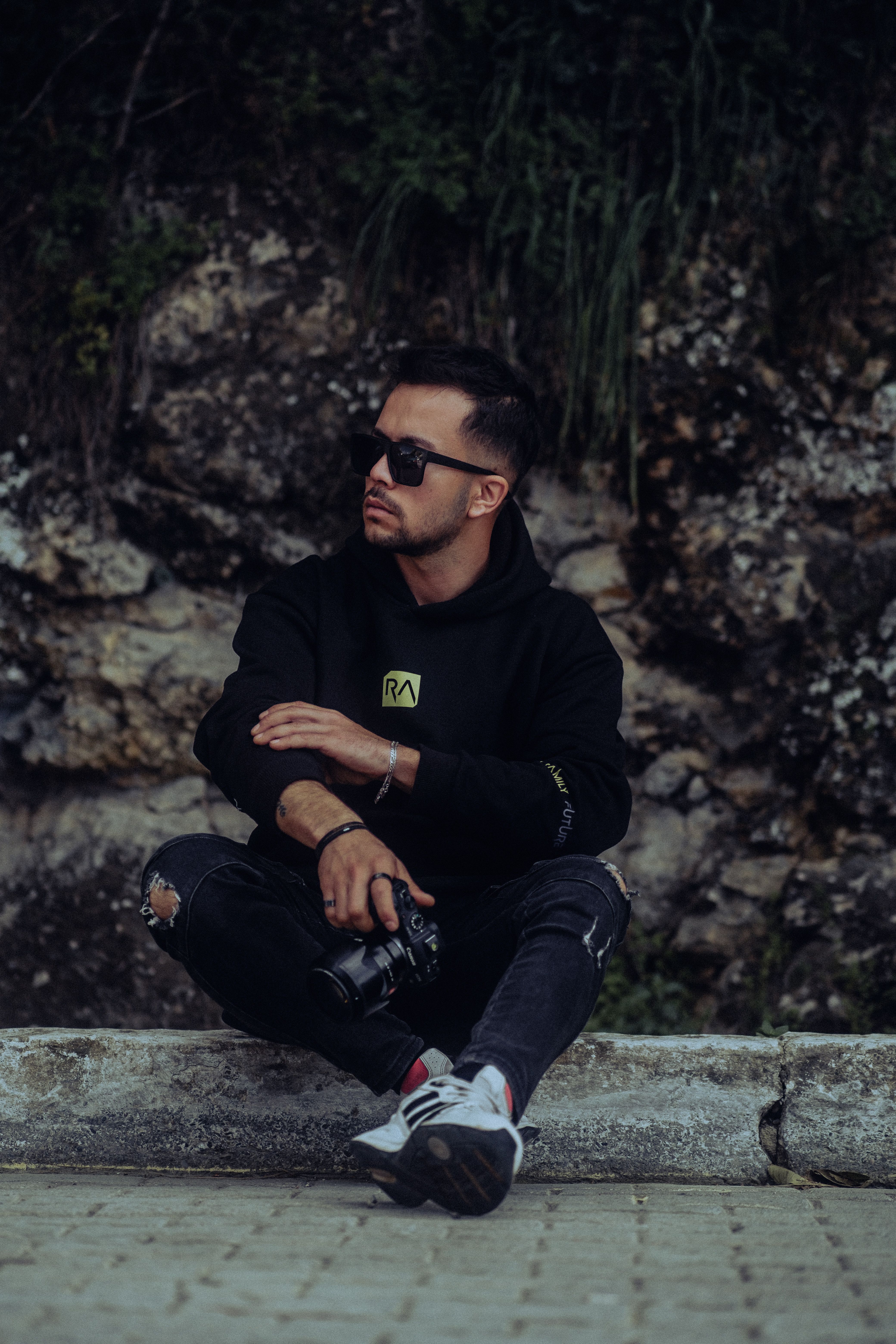 Caucasian man in stylish attire sitting with camera outdoors, looking thoughtful.