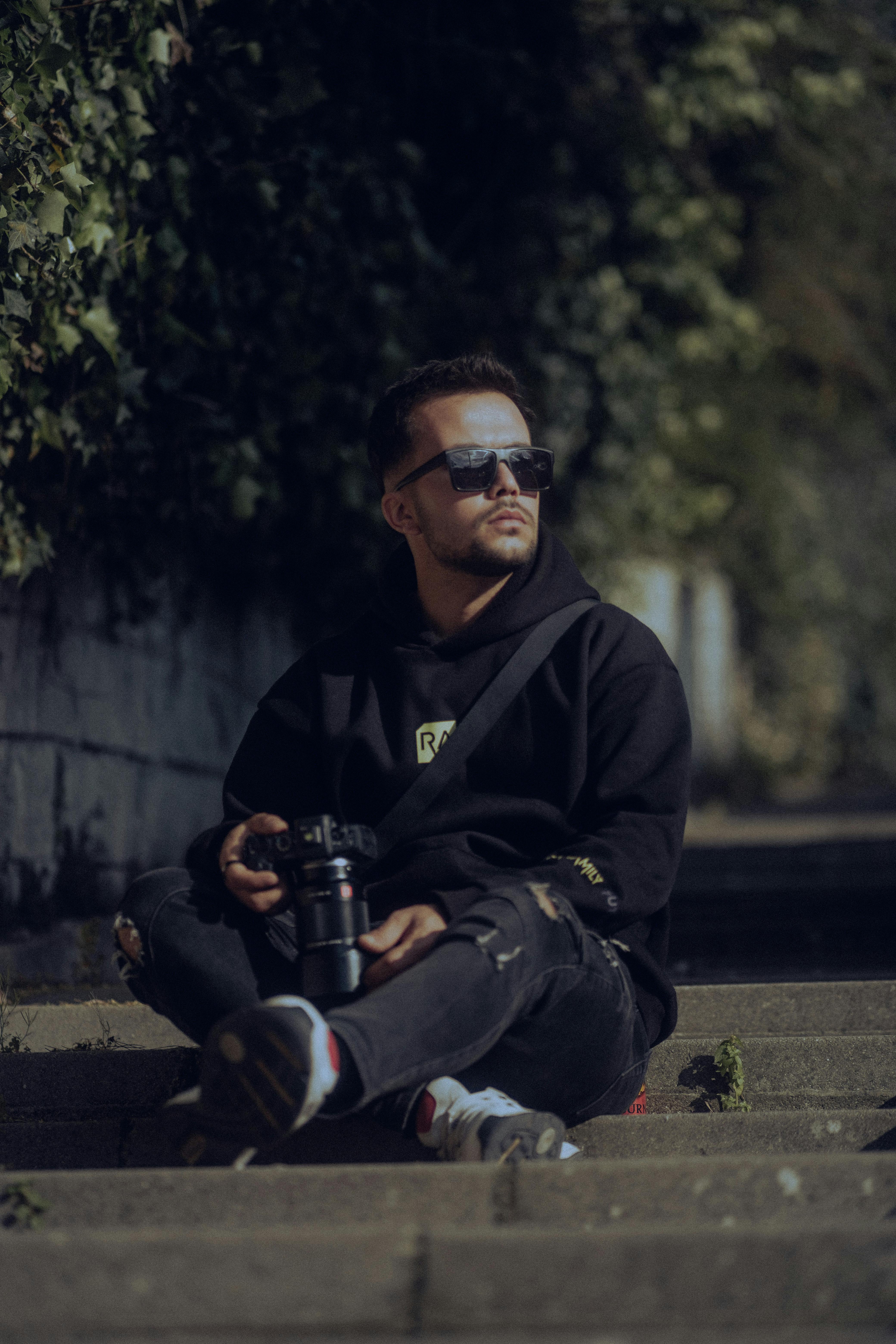 Portrait of a young man holding a camera, sitting on outdoor stairs, wearing sunglasses.