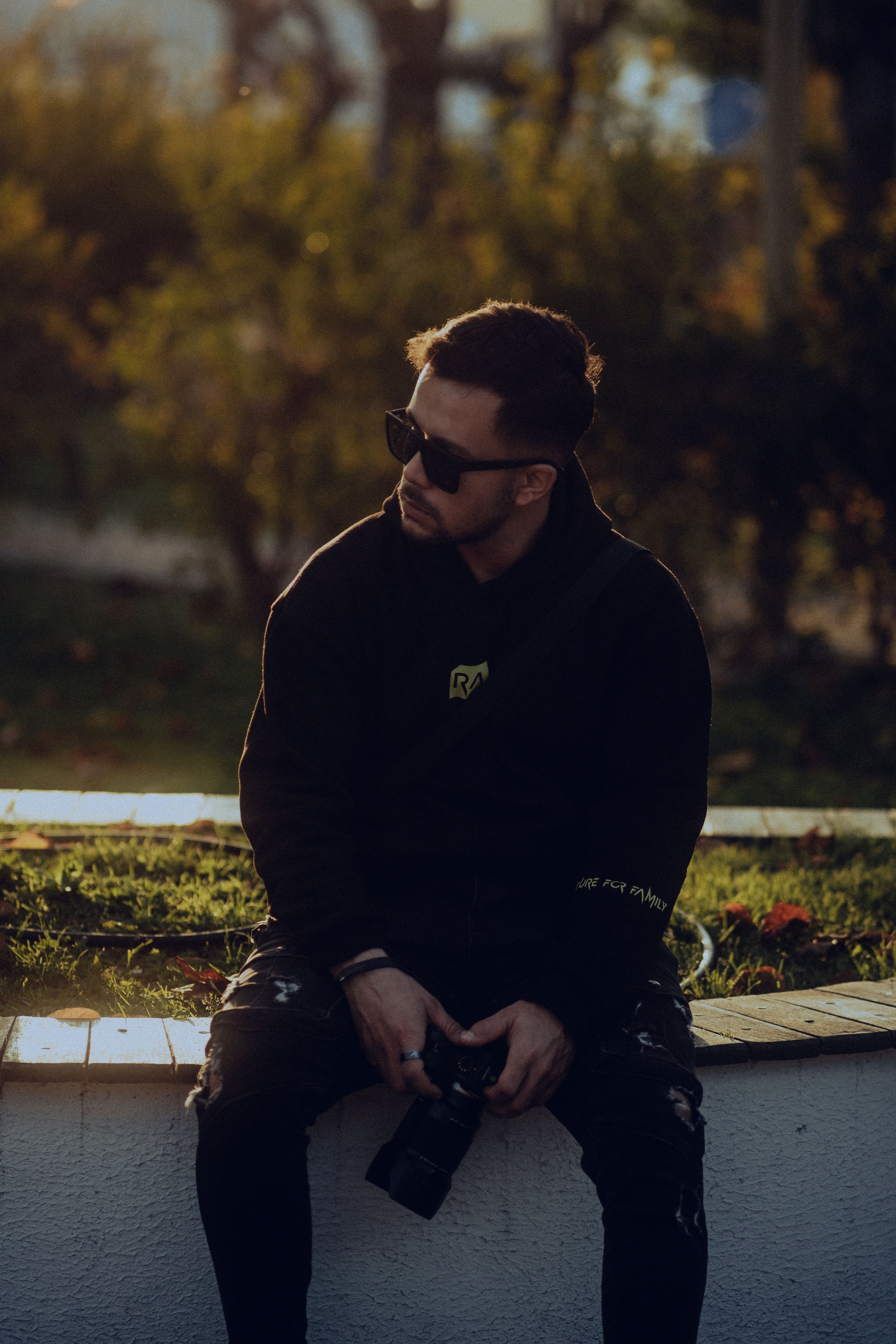 Portrait of a young man in sunglasses sitting outdoors holding a camera during sunset.