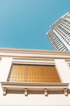 From below window with bars of modern residential building located against skyscraper and blue cloudless sky