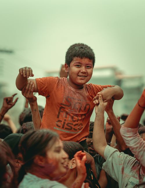 Free Happy child celebrating Holi, lifted by a crowd outdoors, smiling with vibrant colors. Stock Photo