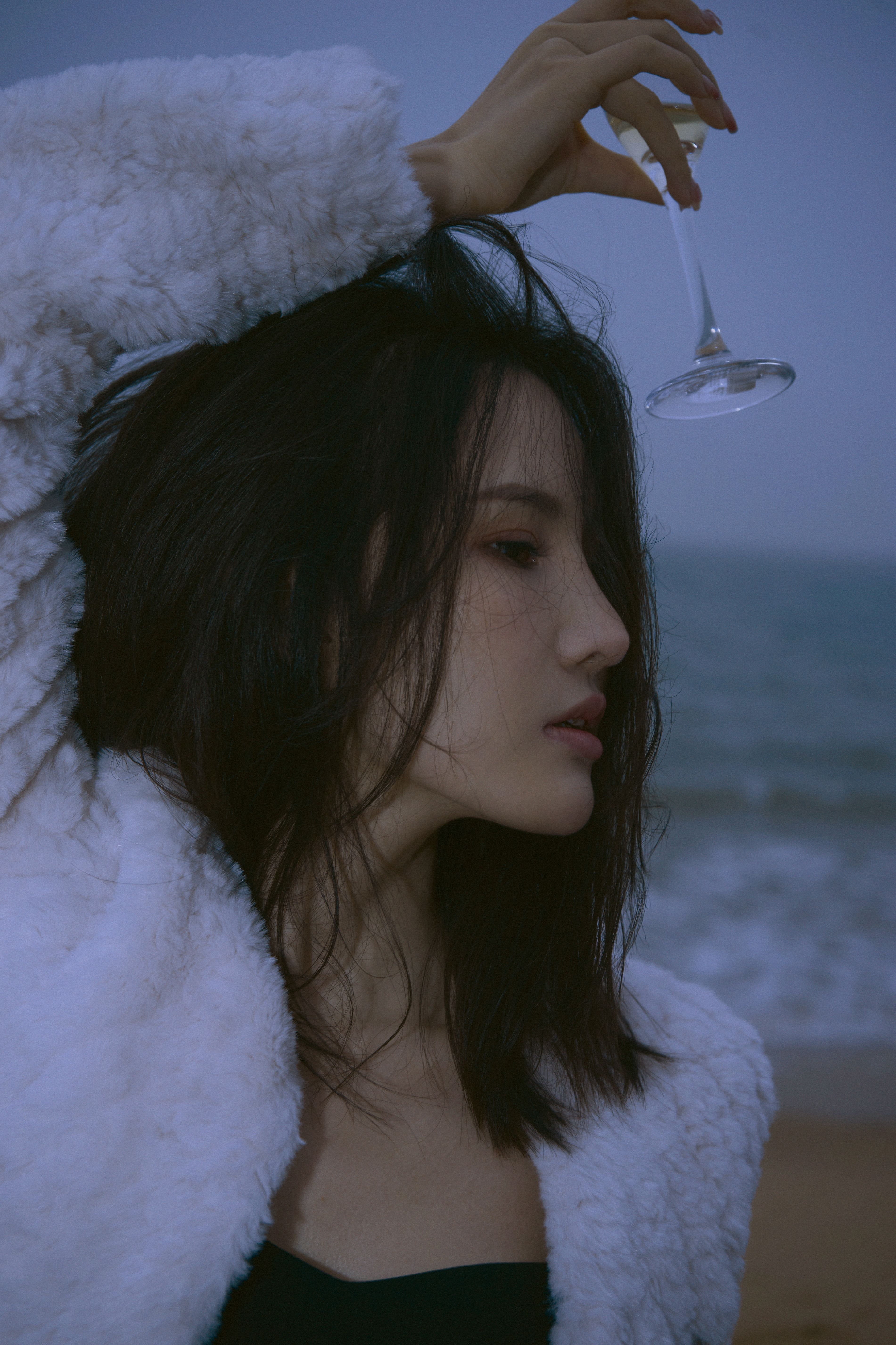 Woman in fashion-forward pose with a glass, standing by the sea wearing a white fur coat.