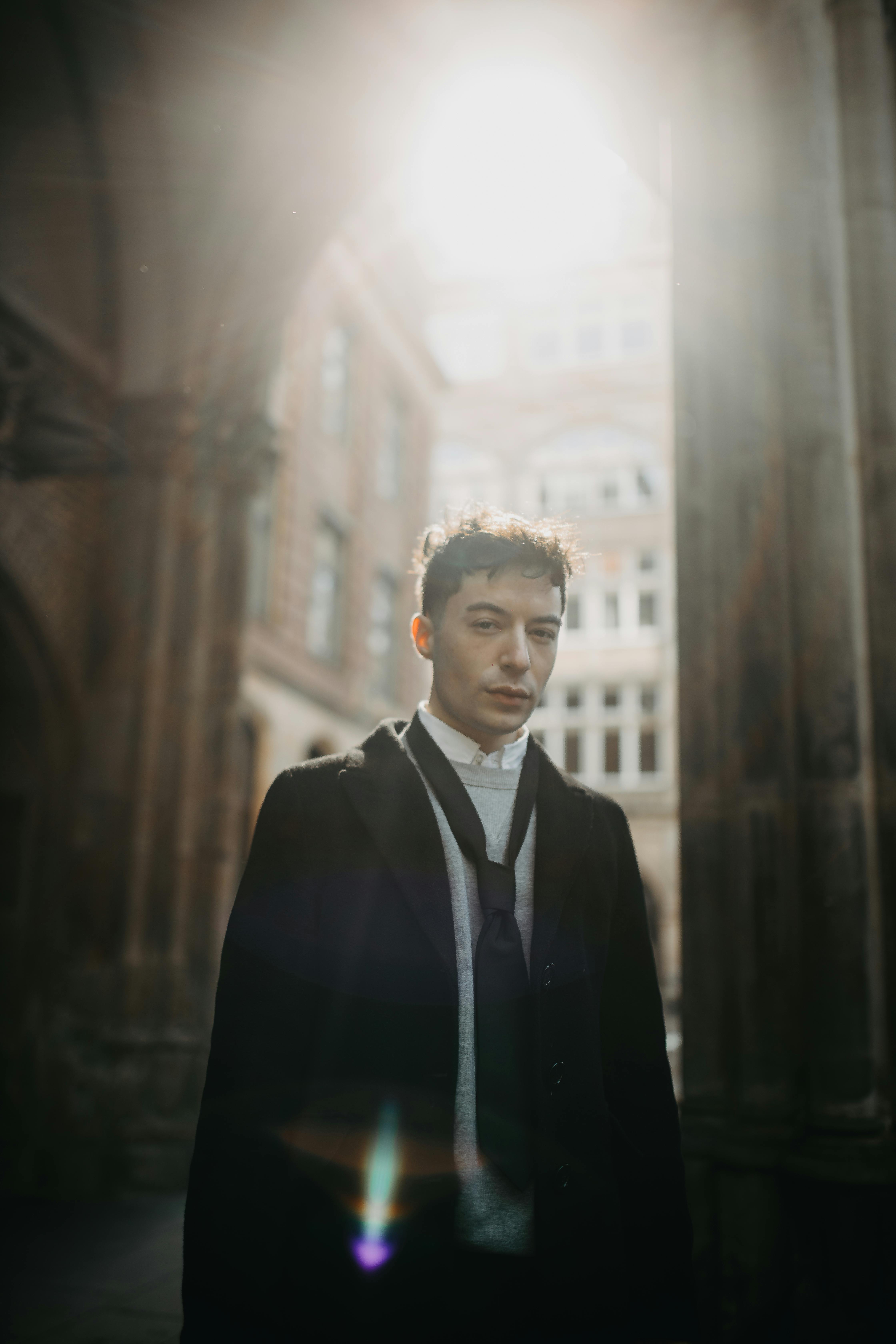 Stylish man in black jacket stands in sunlight against architectural backdrop.