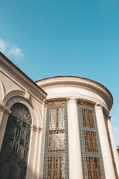 Low angle exterior of white mansion with classic arched and rectangular windows behind ornamental bars against blue sky