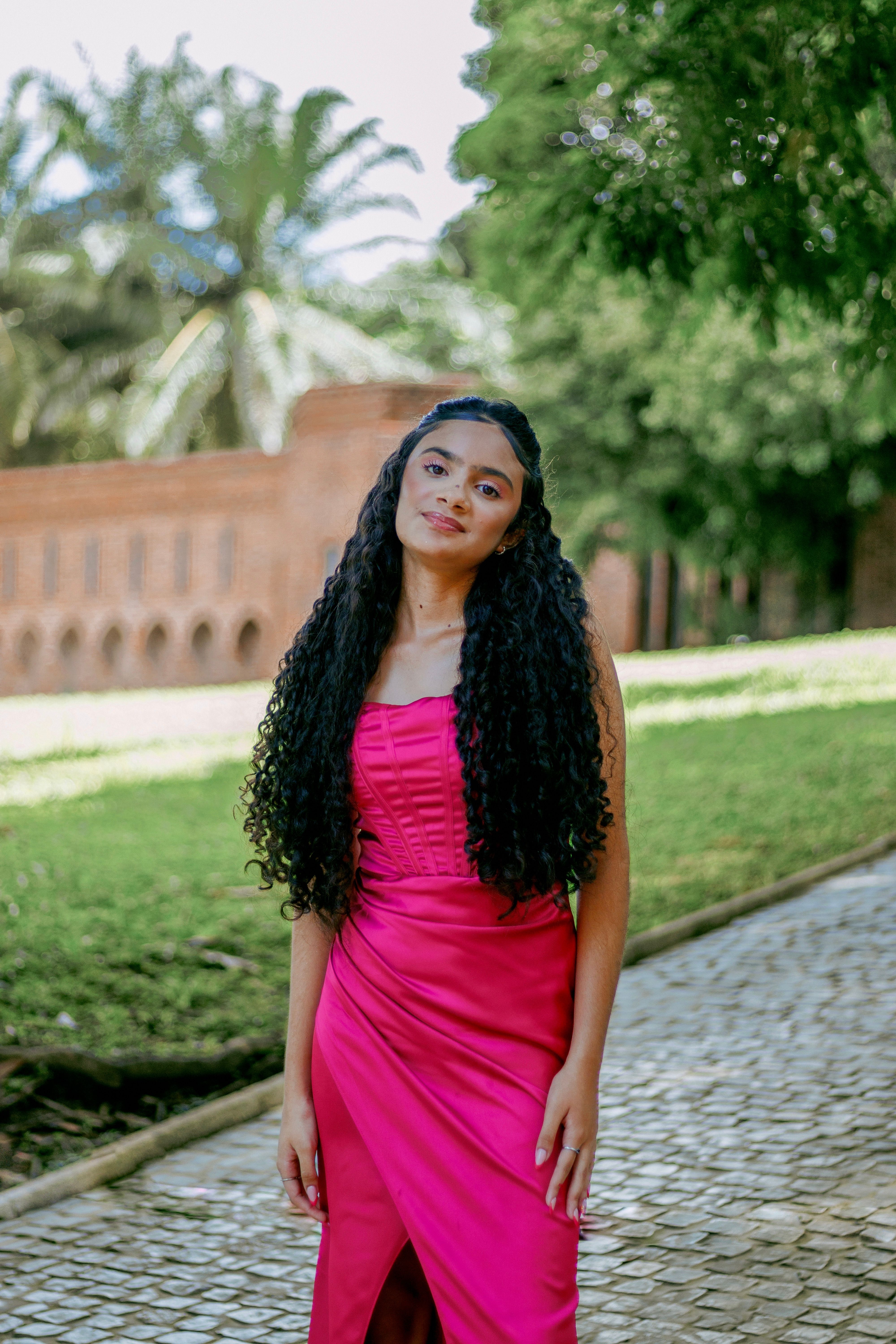 Elegant woman in a vivid pink dress poses outdoors, showcasing fashion and natural beauty.