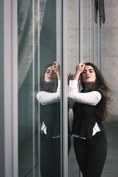 Elegant woman posing by a glass building, capturing urban style and reflection.