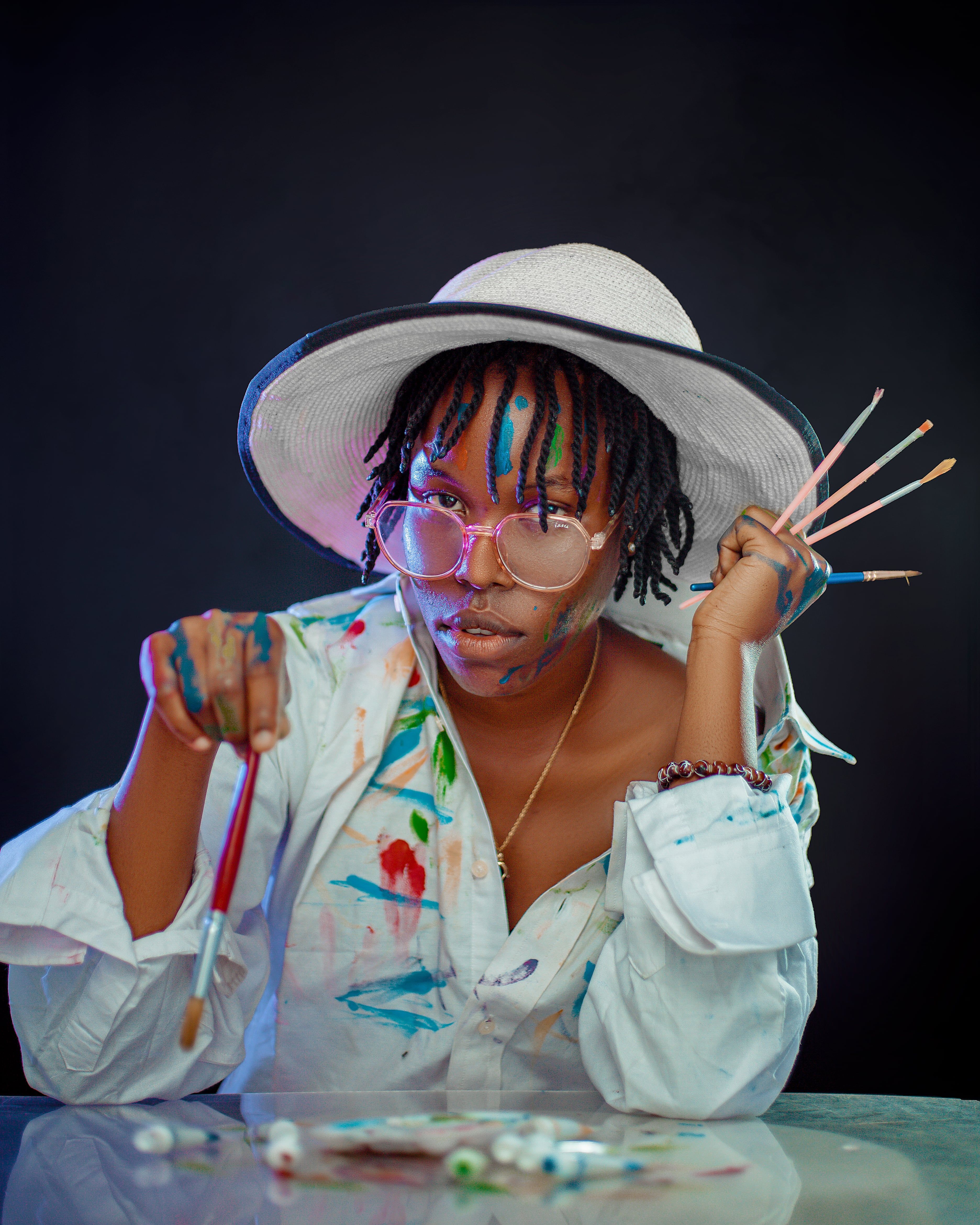 A creative adult artist wearing a hat and eyeglasses, holding paintbrushes with colorful paints.