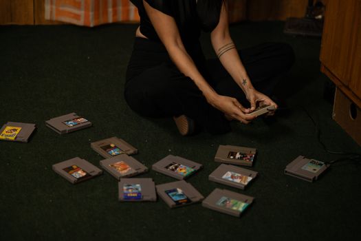 Adult woman sitting cross-legged, holding a vintage game controller, surrounded by retro game cartridges.