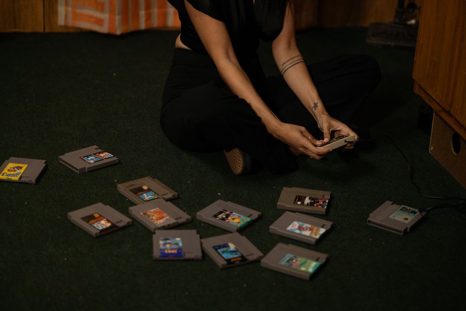 Adult woman sitting cross-legged, holding a vintage game controller, surrounded by retro game cartridges.