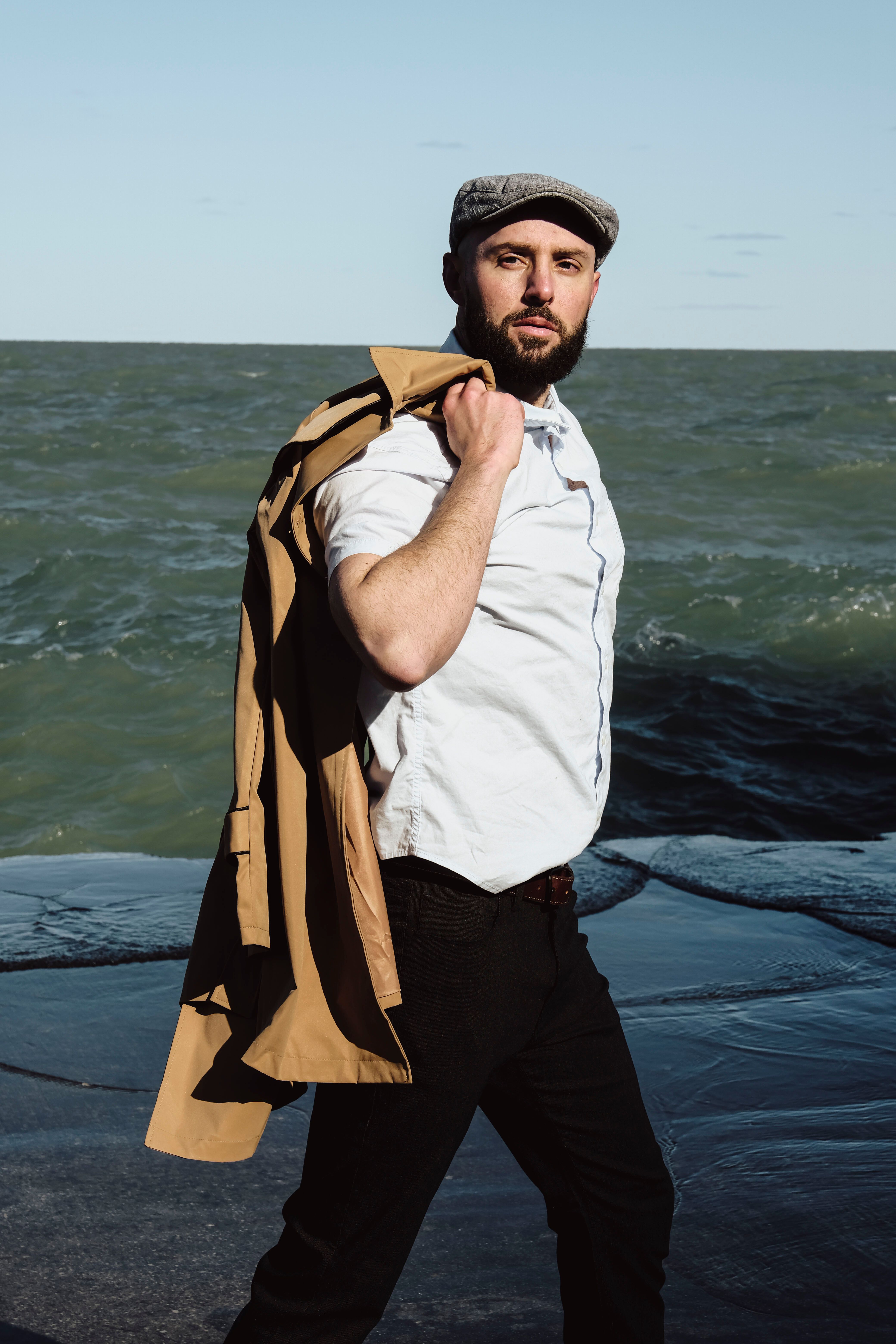Stylish man with coat over shoulder walks along the seaside, showcasing modern fashion.