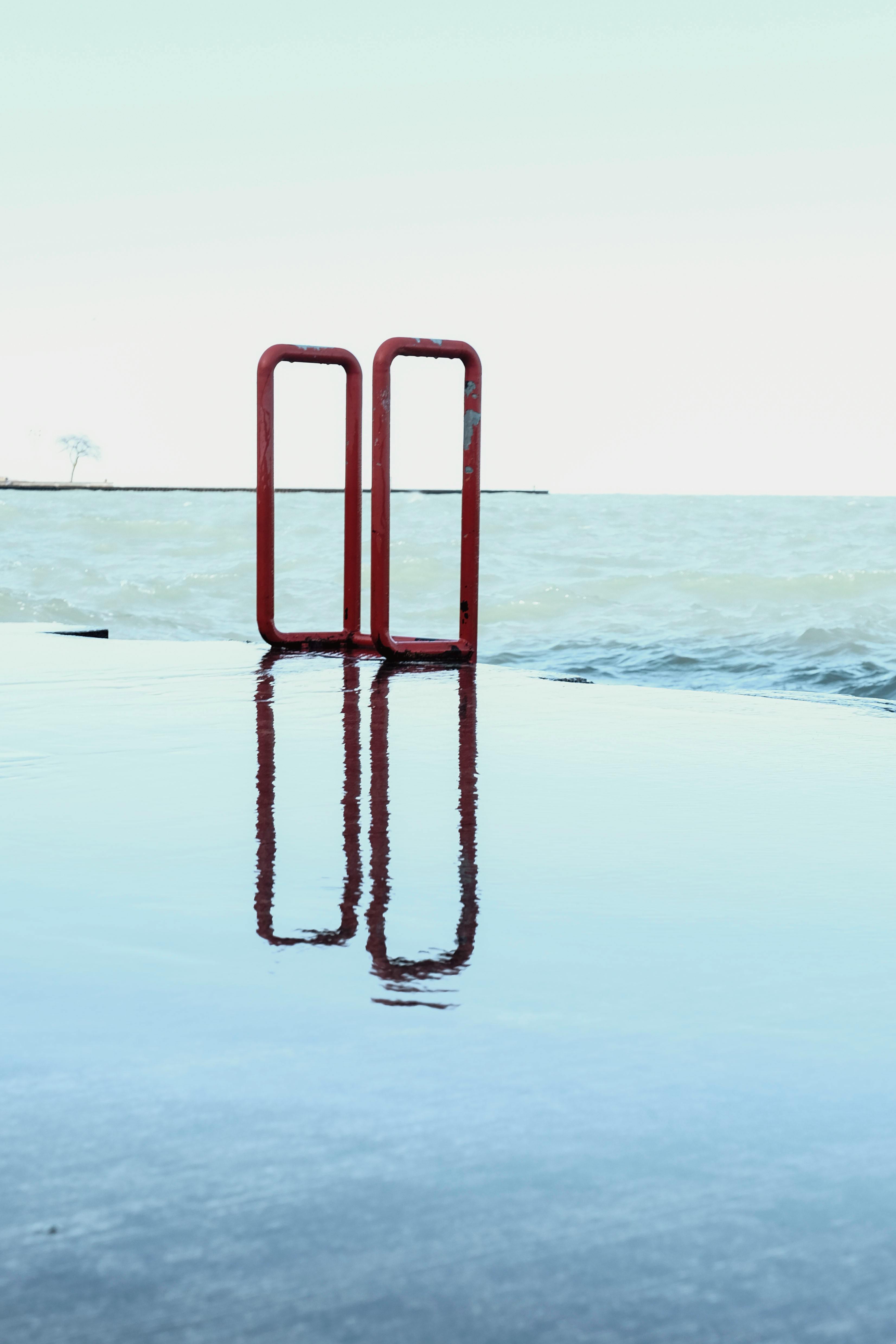 Red metal handrails reflecting in calm water by the seashore on a clear day.