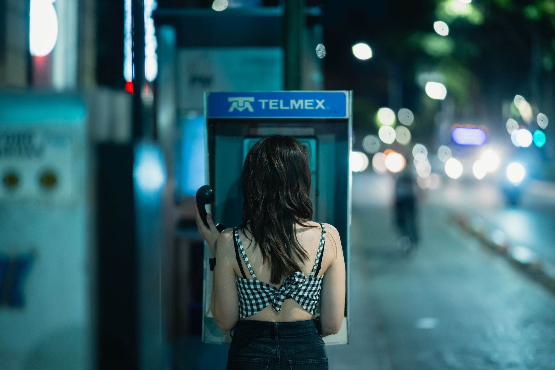 A woman stands at a TELMEX payphone booth on a city street at night with blurred lights.