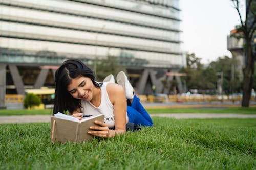 Smiling Woman Lying Down on Grass at Park and Writing in Notebook