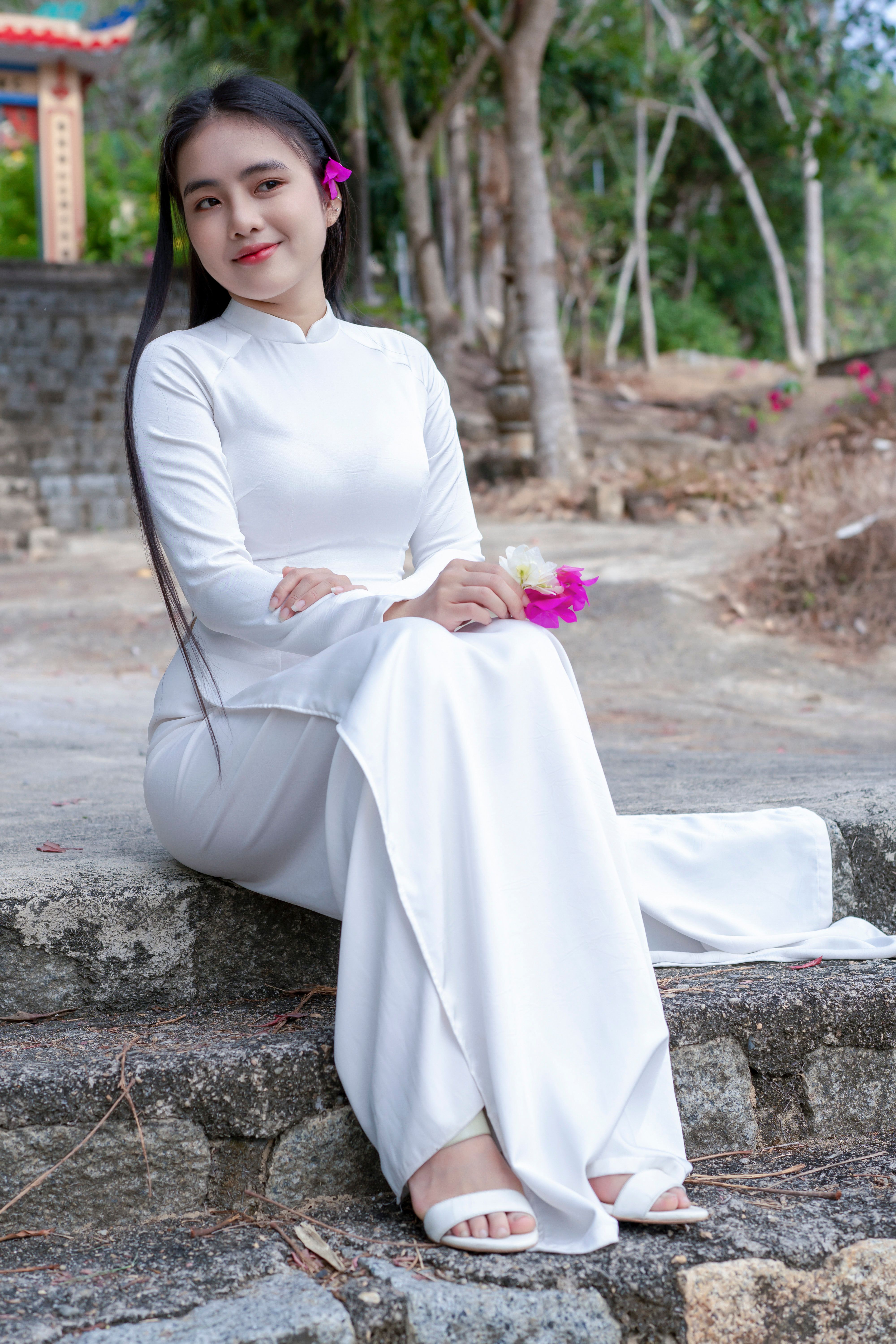 A graceful woman in a white Ao Dai sitting outdoors, showcasing traditional Asian attire.