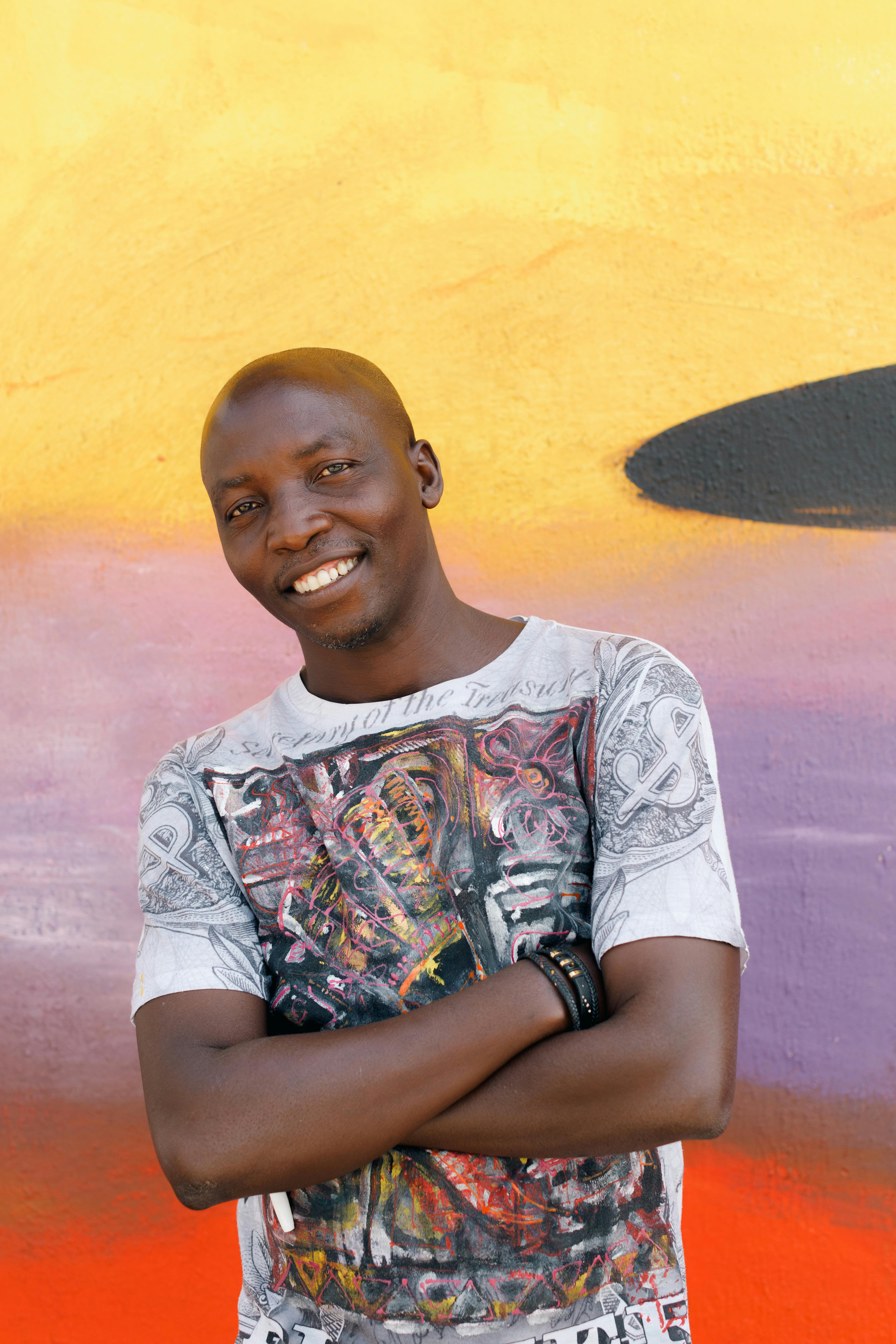 Happy man in Lagos standing by a colorful mural, arms crossed, wearing a graphic t-shirt.