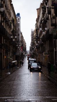 A rainy urban street scene featuring cobblestone roads, cars, and towering buildings on either side.