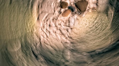 Abstract image of pebbles creating a swirling pattern in a riverbed, depicting earth tones and tranquil motion.