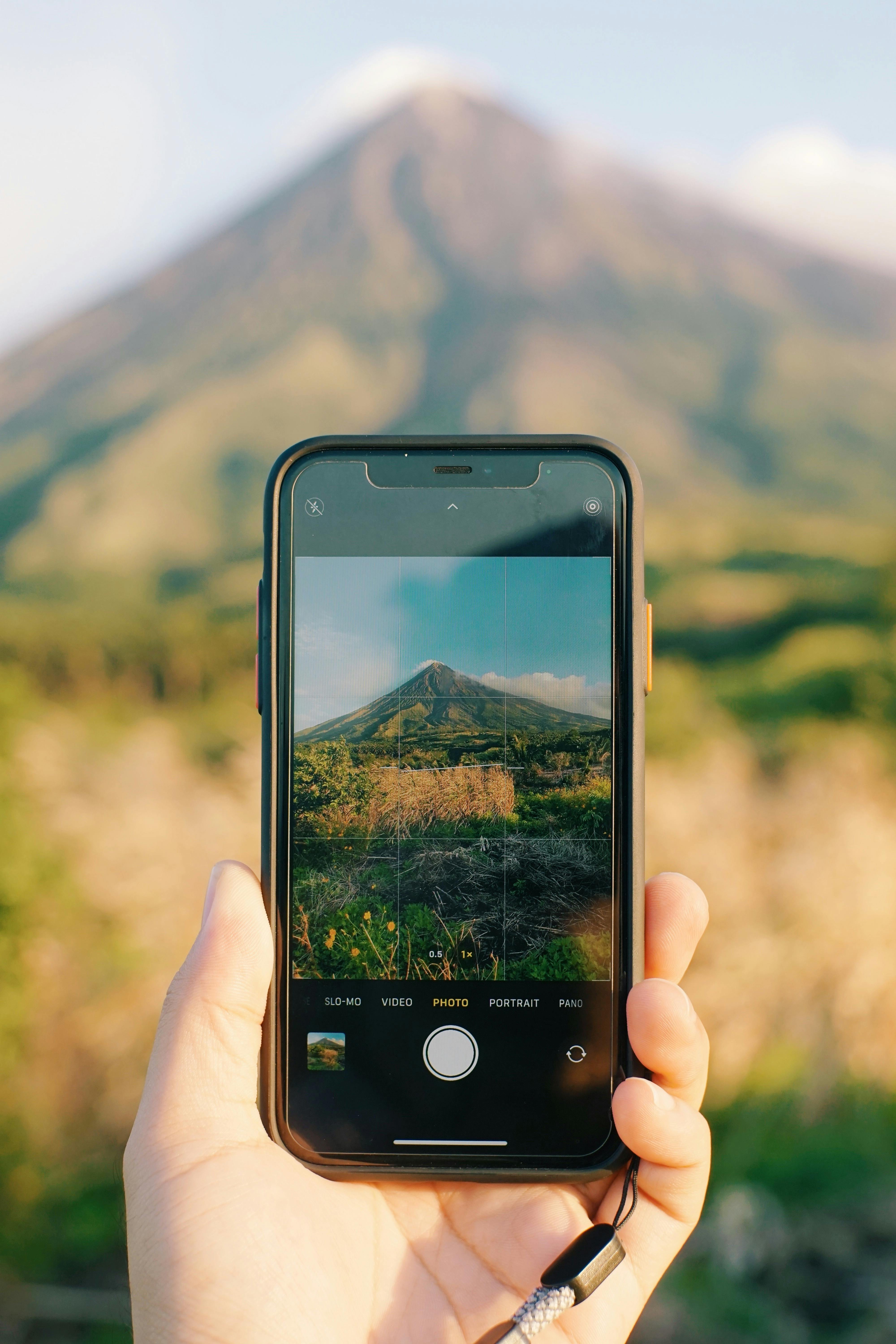 A hand holding a smartphone capturing a picturesque volcano on a sunny day.