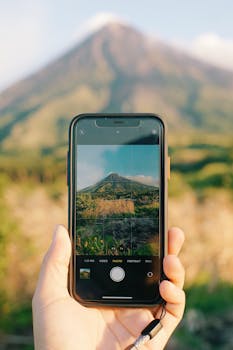 A hand holding a smartphone capturing a picturesque volcano on a sunny day.