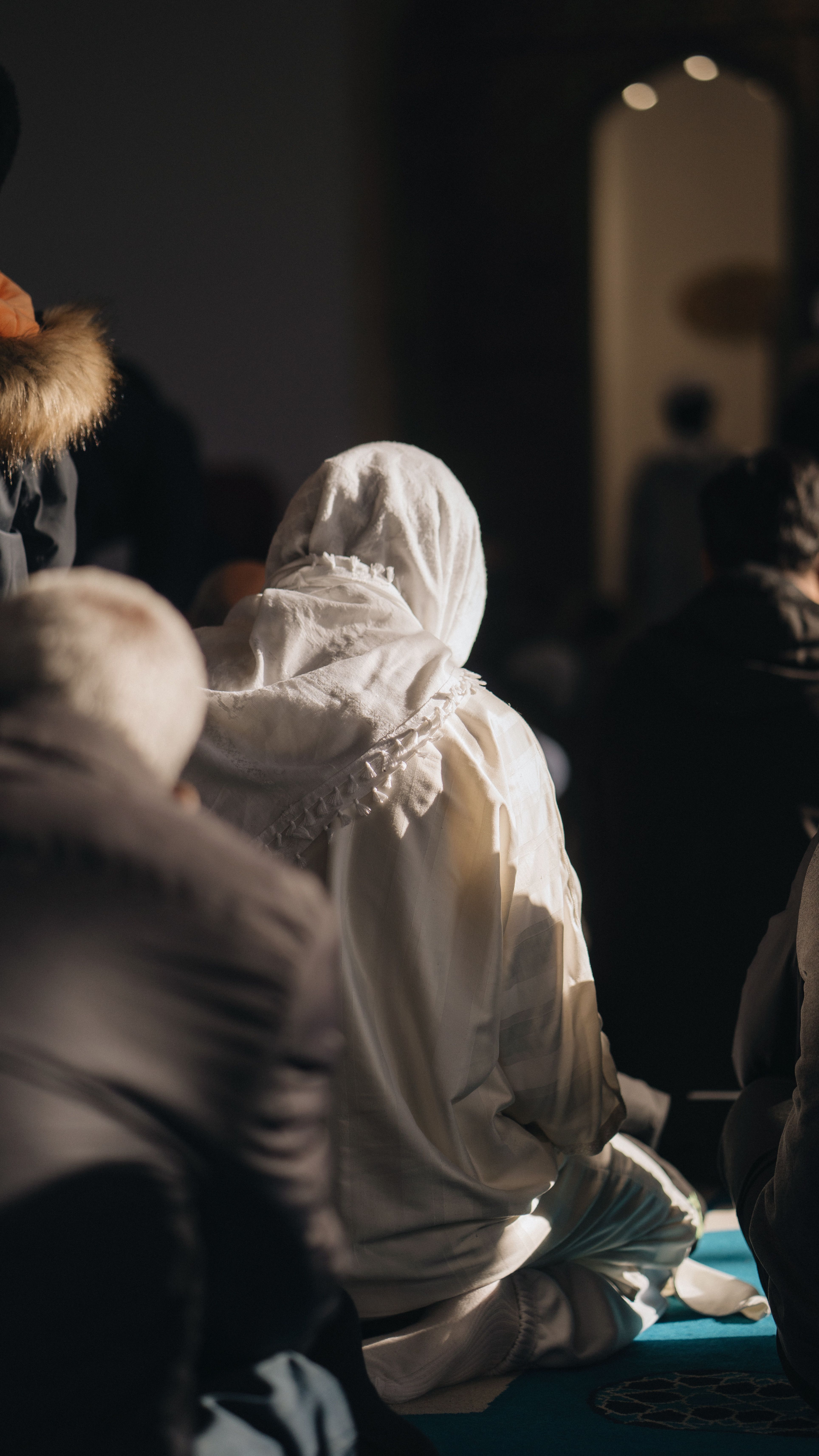 A Muslim woman in white prays in a mosque with sunlight illuminating her among others.