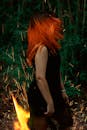 篝火旁的森林中，站着一位身穿黑色连衣裙、一头火红长发的女子
