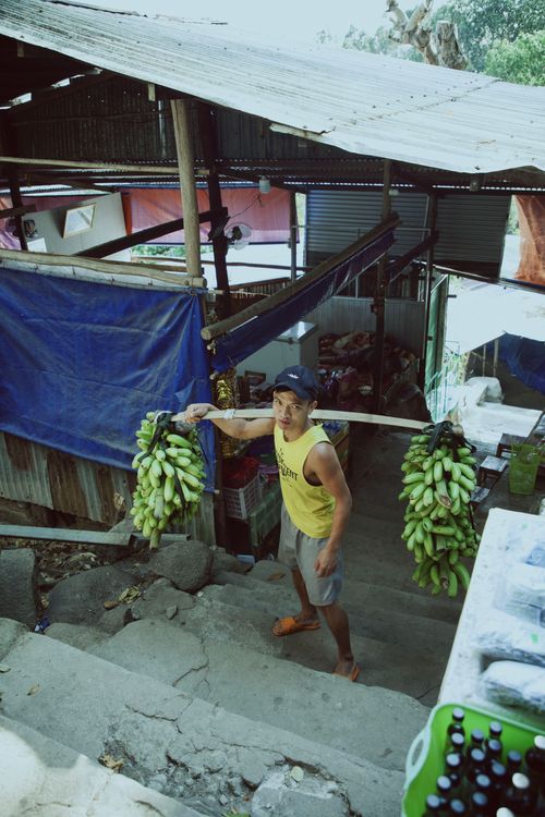 Free A man in a yellow tank top carries bananas in an urban street market setting, showcasing daily life. Stock Photo
