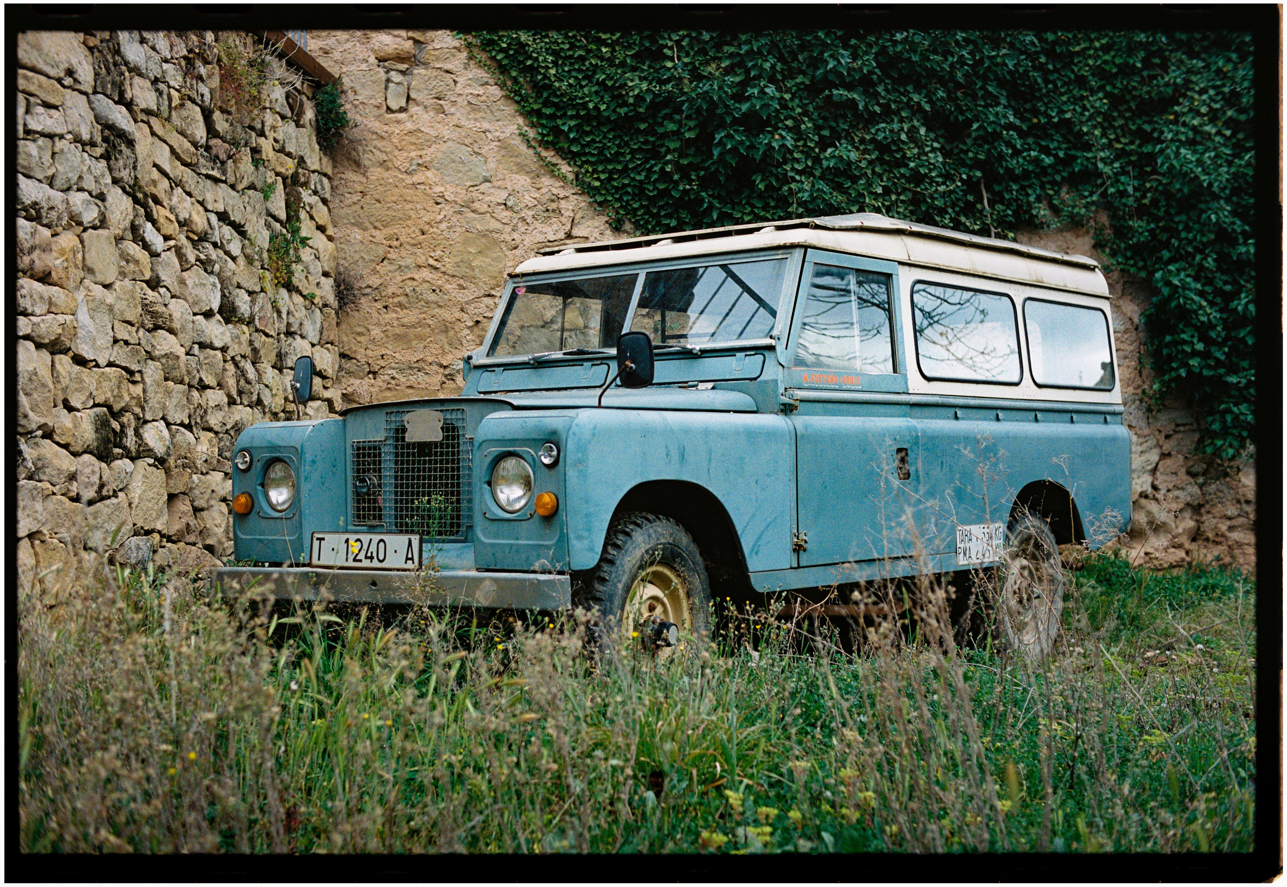 Classic blue Land Rover parked beside an ivy-covered stone wall in a rural setting.