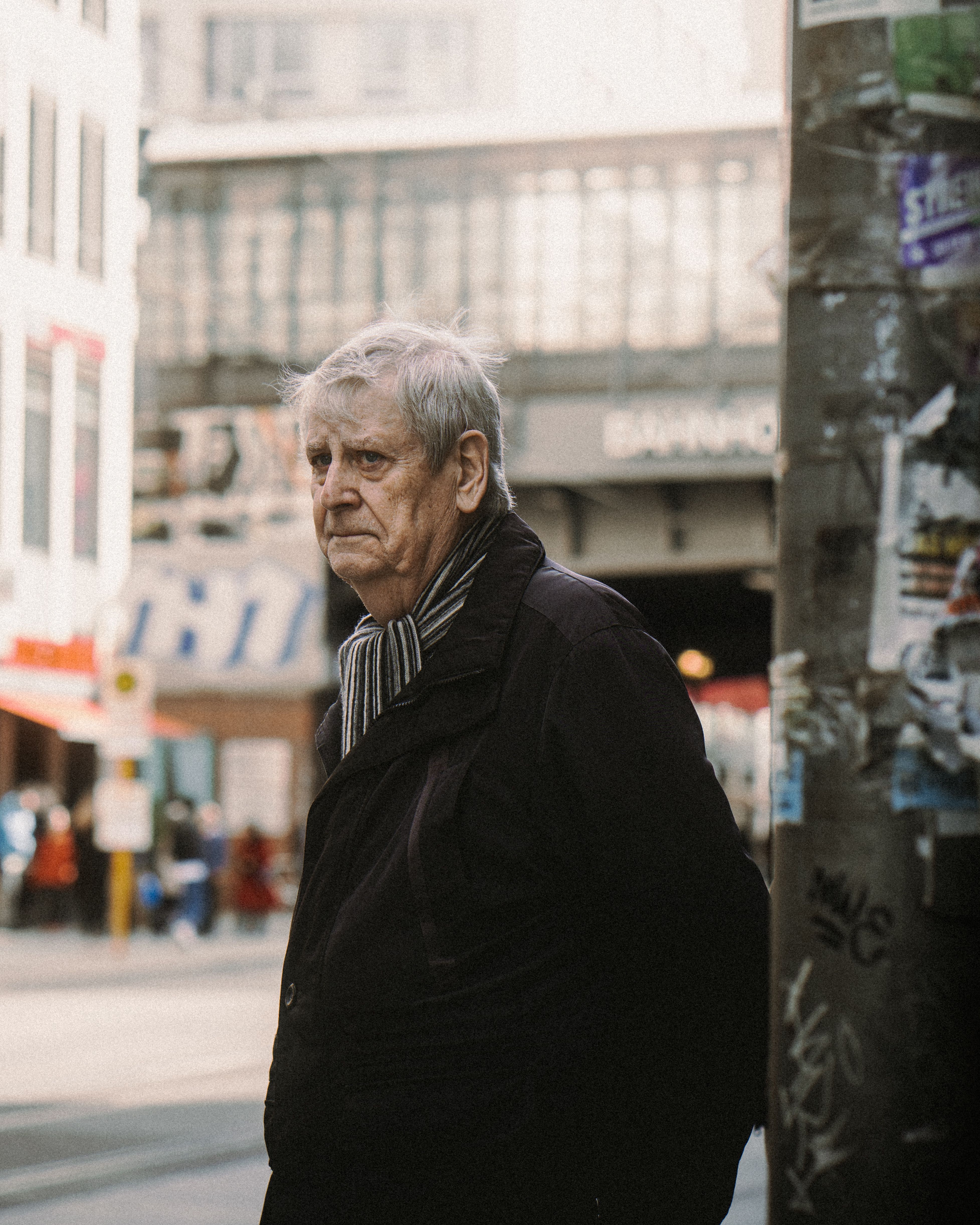 An elderly man with gray hair stands in an urban Berlin setting, wearing a black jacket.