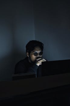 An Asian man with glasses sits in a dimly lit room, focused on his computer work.