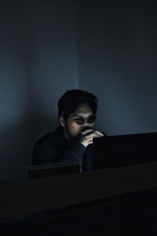 Free An Asian man with glasses sits in a dimly lit room, focused on his computer work. Stock Photo