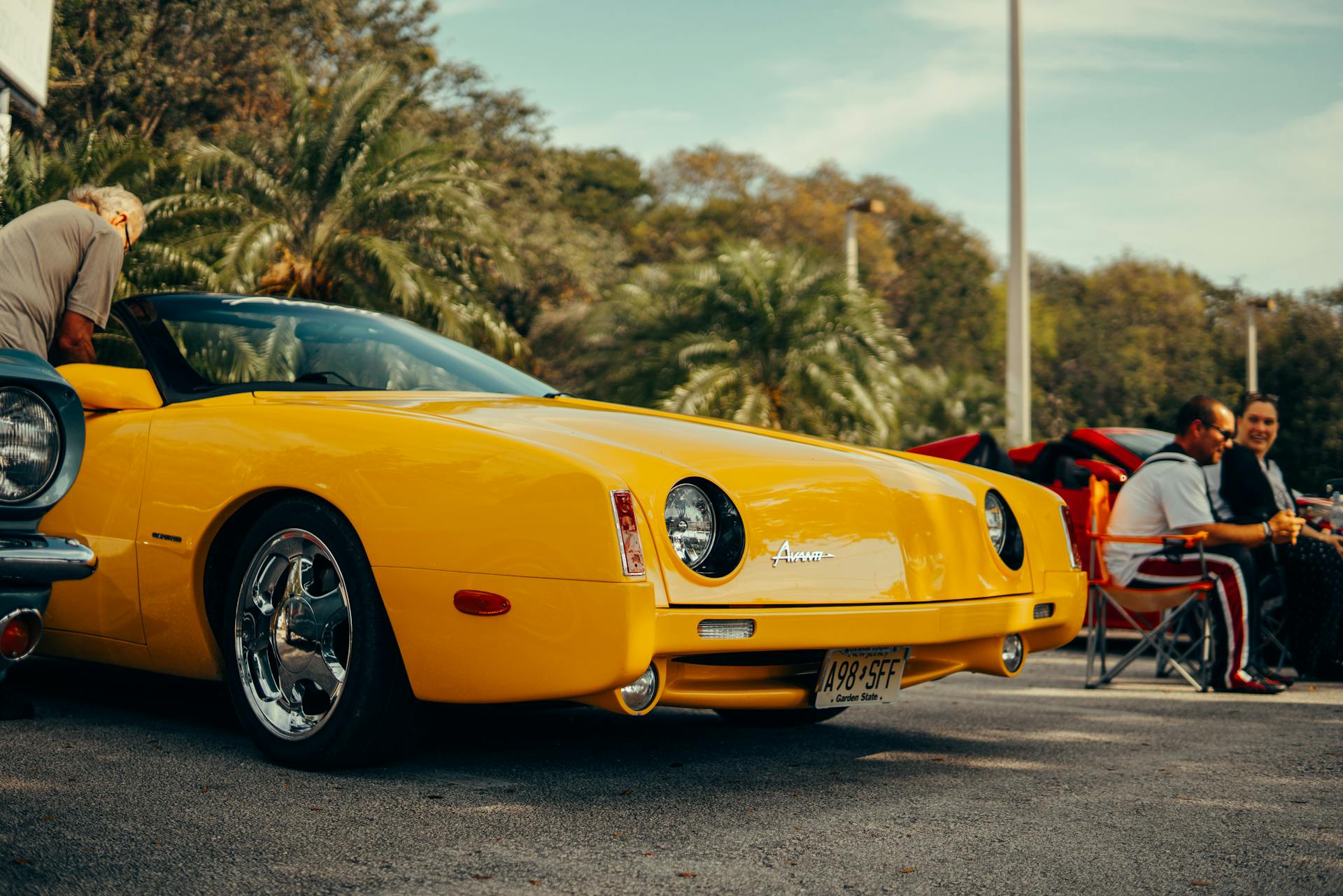 A classic yellow Avanti convertible parked outdoors with people in the background.