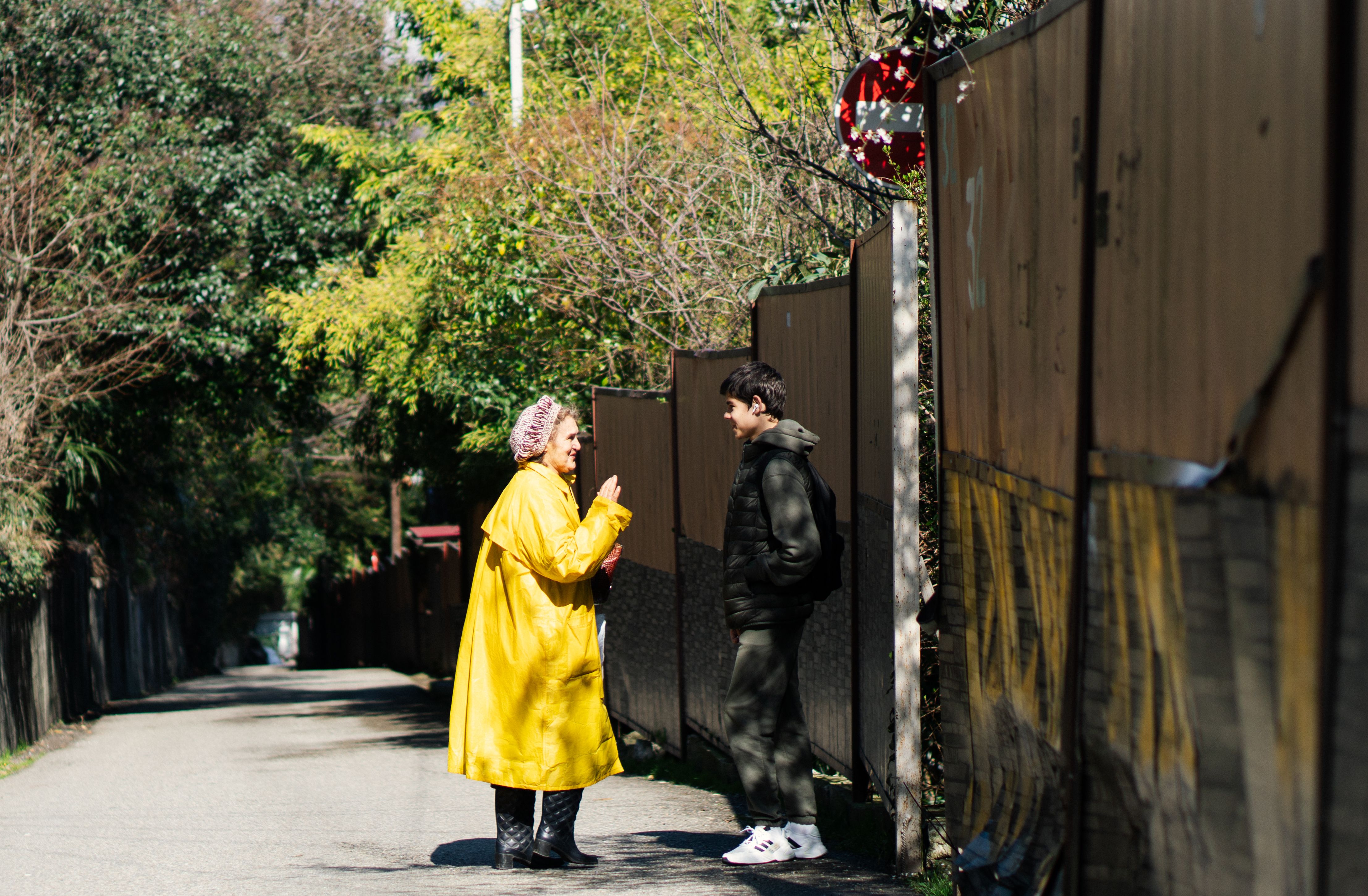 A woman in a yellow coat talks to a teenager on a city street in daylight.
