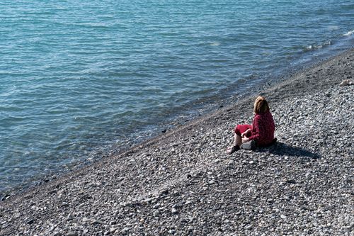 Free Woman in red sits on a rocky beach by the blue sea under bright sunlight. Stock Photo