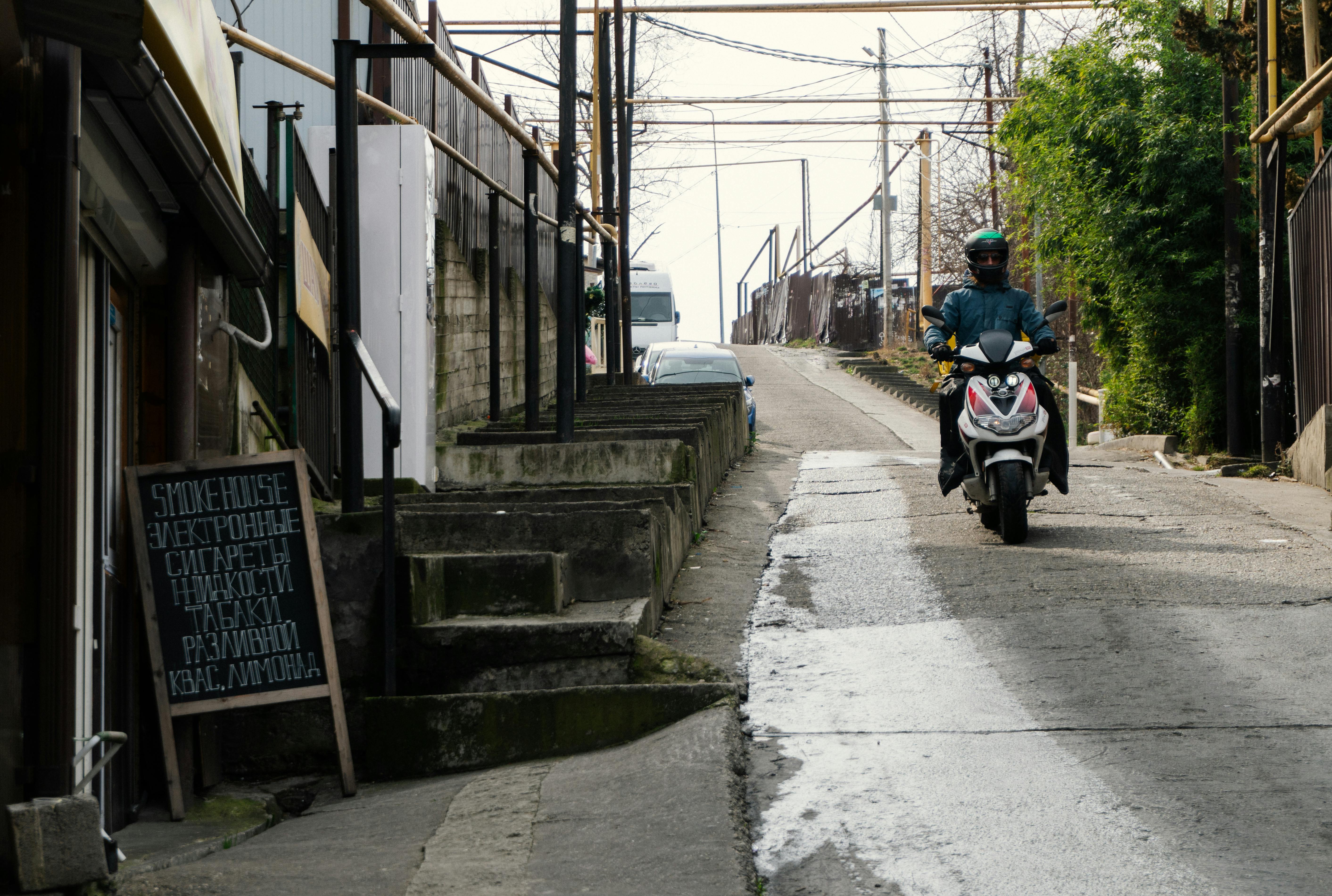 Motorcyclist rides a scooter up a narrow urban street with signs and buildings.