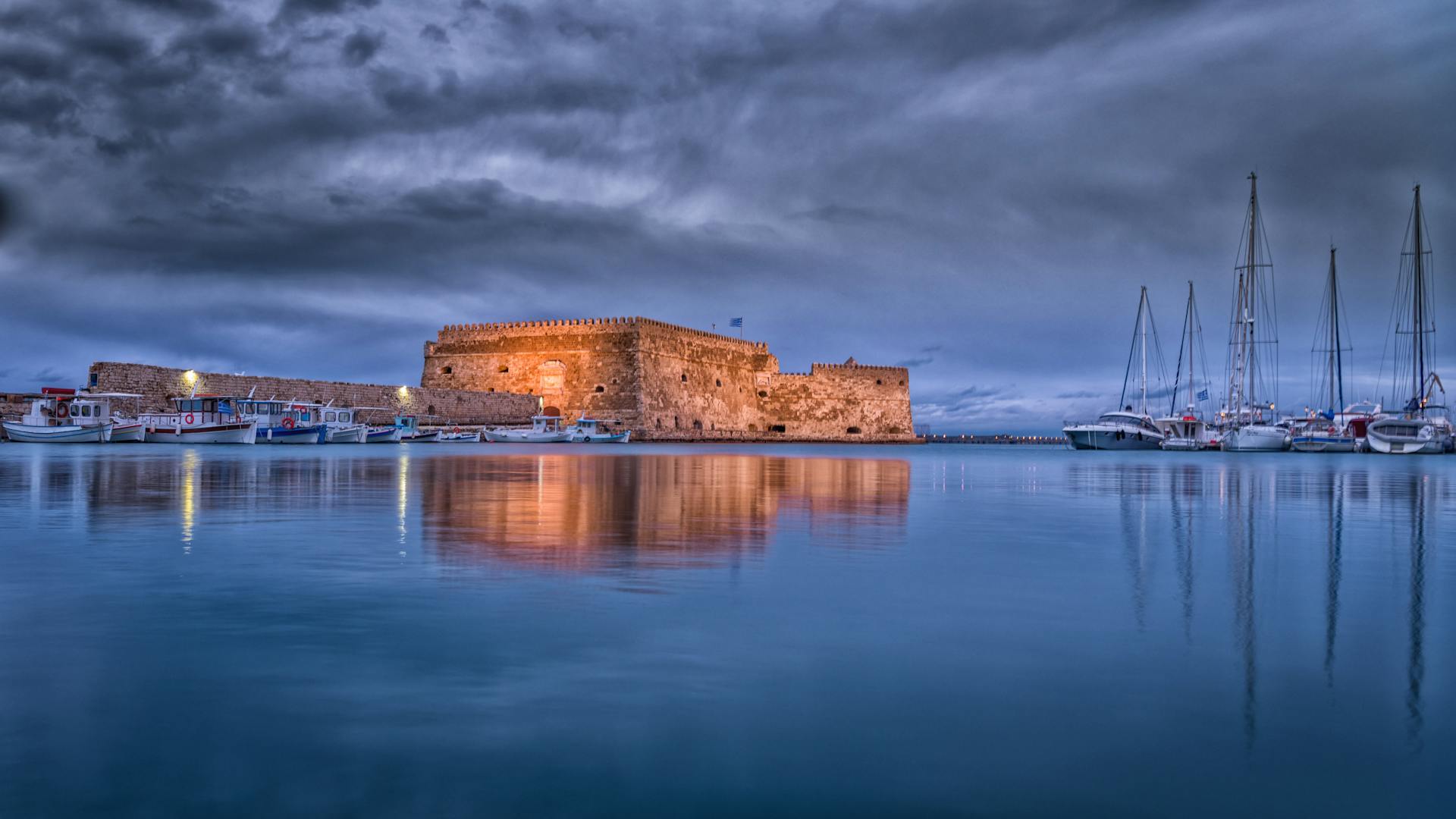 Majestic view of Koules Fortress reflected in calm waters at Heraklion harbor during twilight.