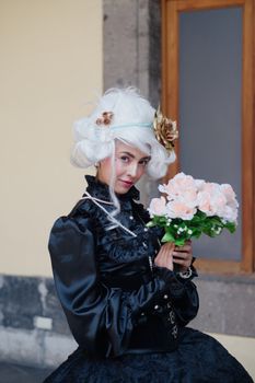 Woman in black attire with white wig holding roses by a window.