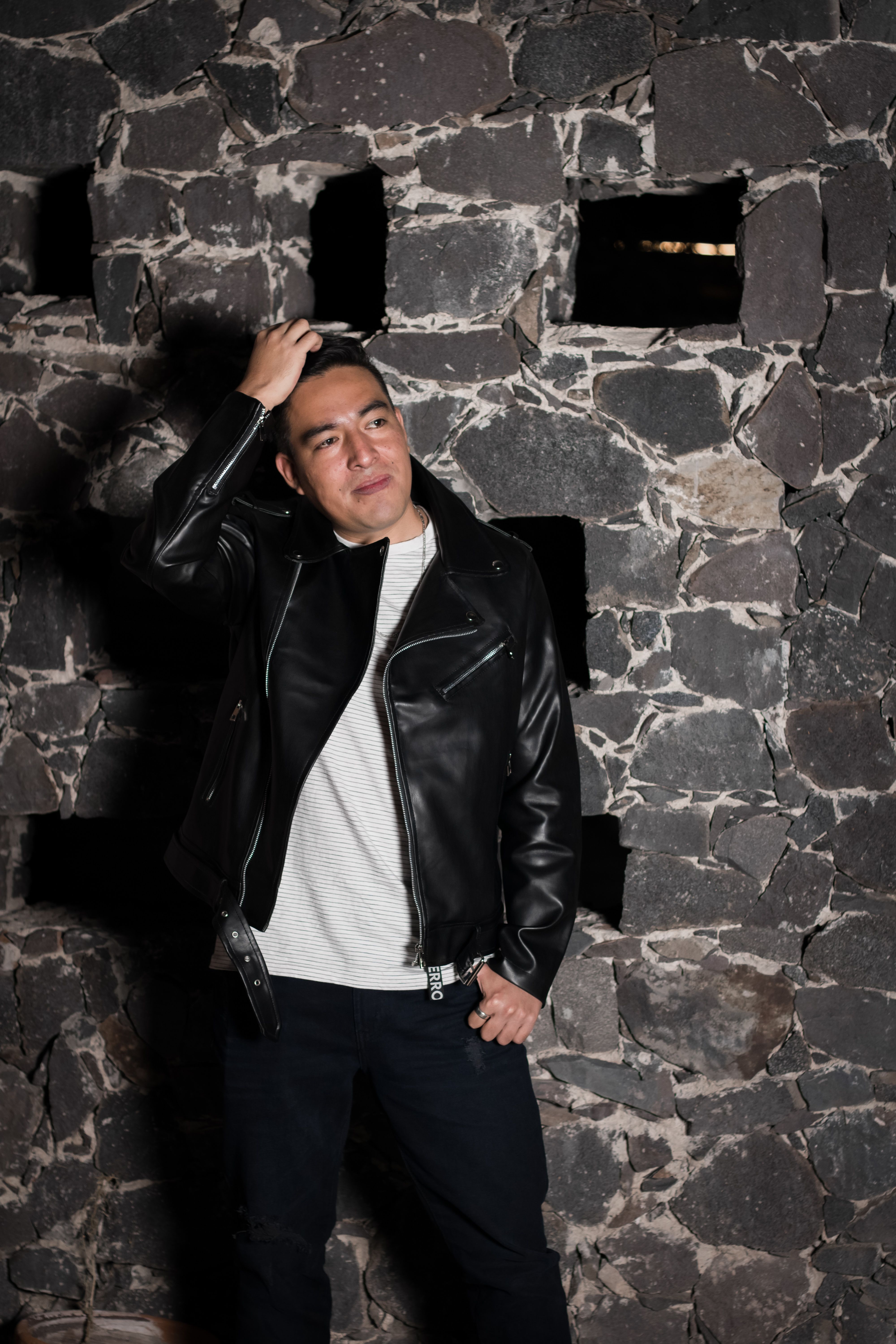 Stylish portrait of a man in black leather jacket posing confidently in front of a rustic stone wall.