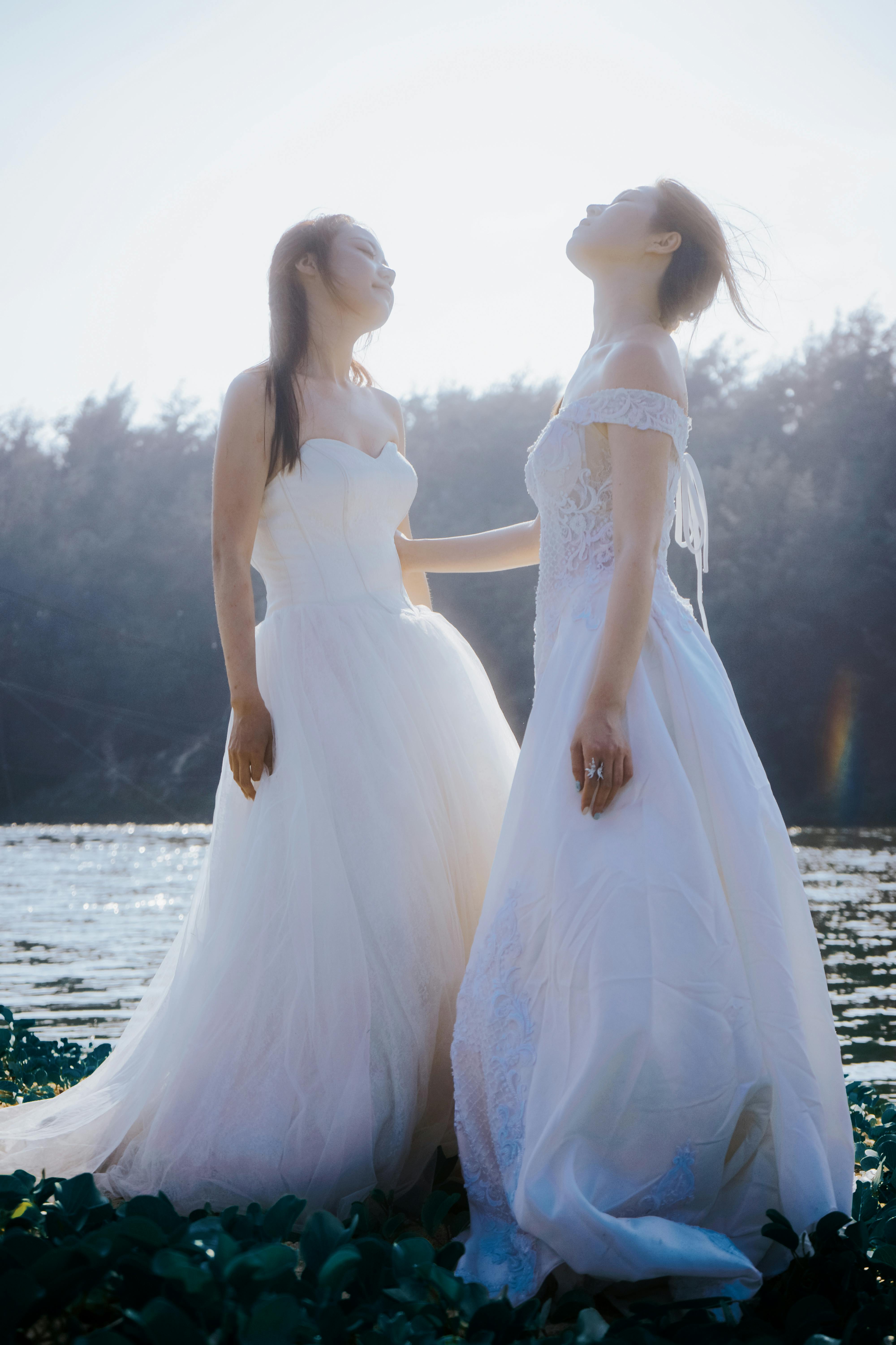 Two brides in beautiful white dresses standing by a sunny lakeside, showcasing elegance and tranquility.