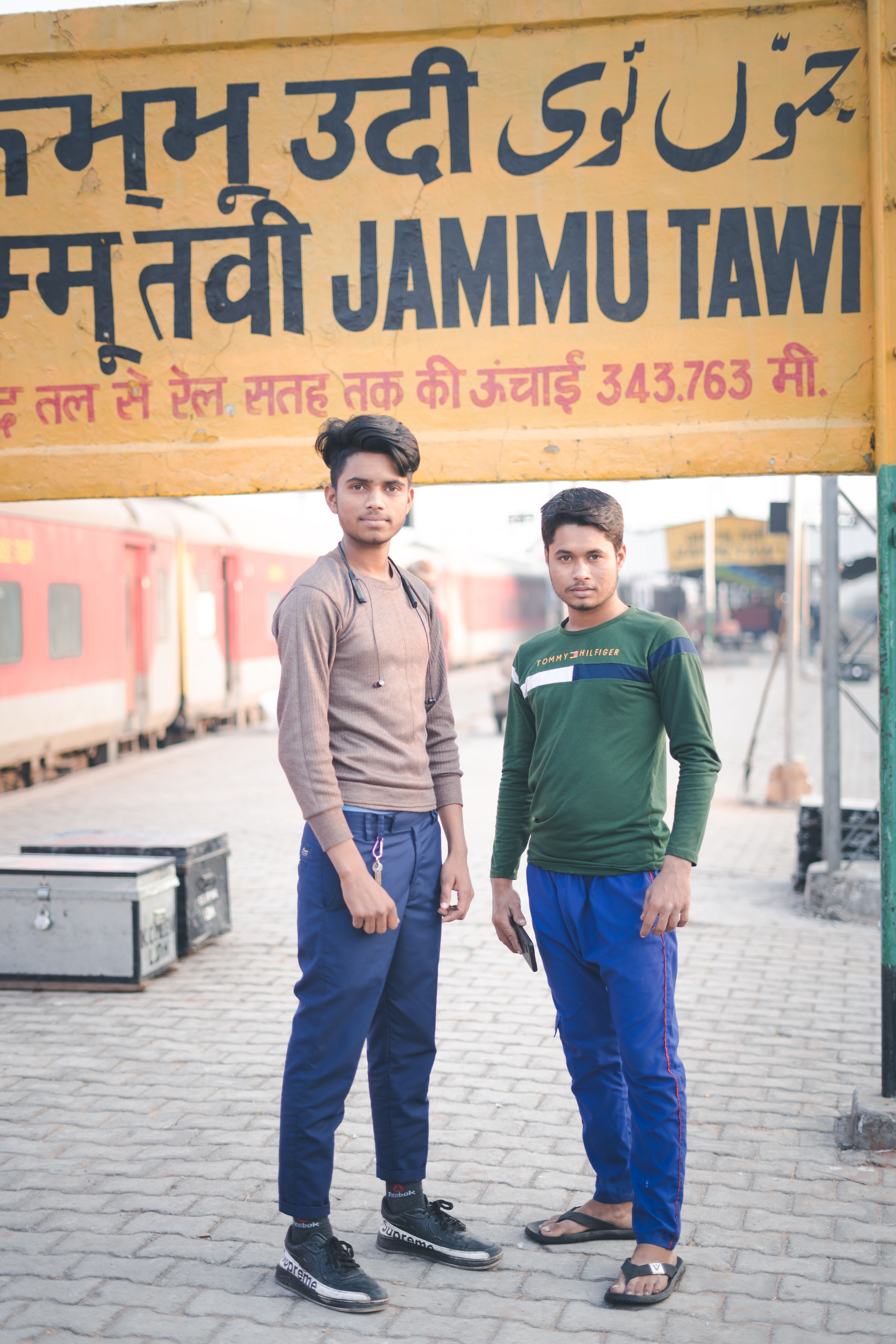 Two young men standing at Jammu Tawi railway station in casual attire.