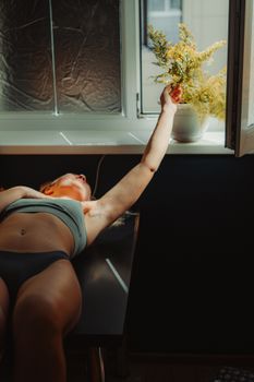 A woman in underwear reaches towards a sunlit potted plant by a window, conveying relaxation and serenity.