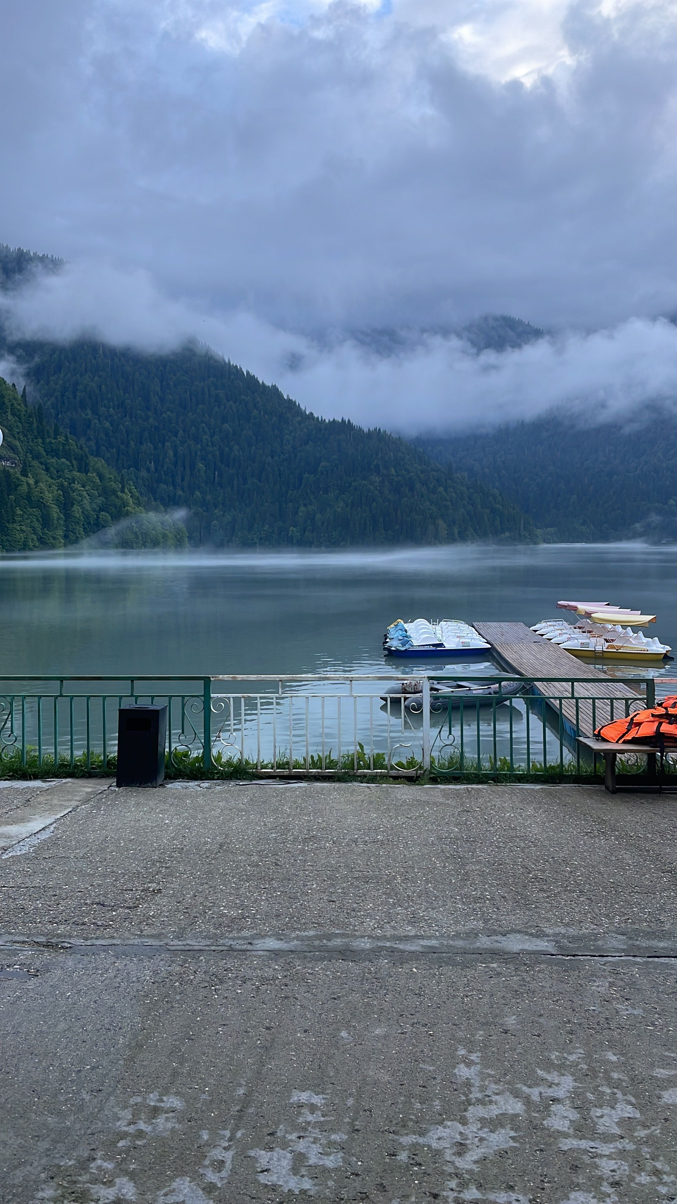 Serene mountain lake with boats, misty forest, and peaceful ambiance under a cloudy sky.
