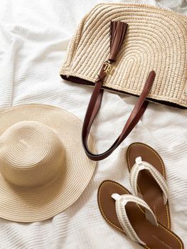 Flat lay of a straw hat, flip flops, and woven bag on a white sheet.